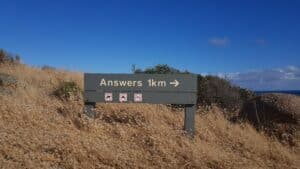 Photo of a sign in a field that reads "Answers 1 km -->" against a blue sky.