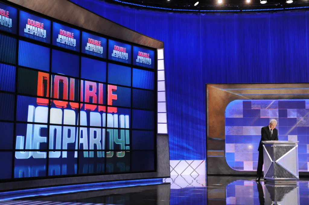 Photo of Alex Trebek on the set of ‘Jeopardy!’ with the Double Jeopardy logo displayed on the game board. 