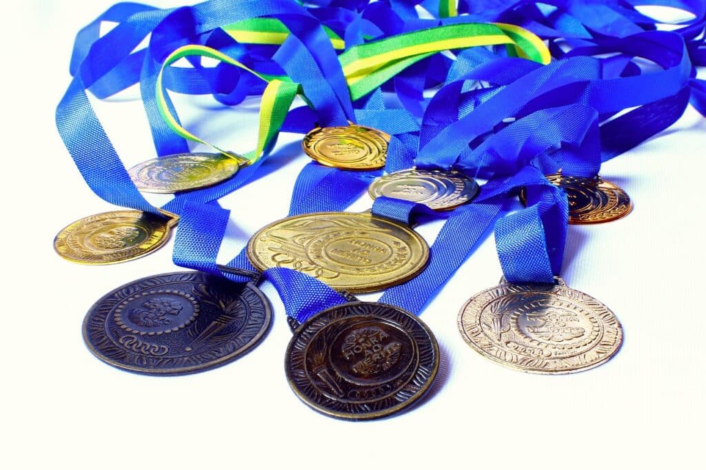 Photo of various gold, silver, and bronze medals with blue and green ribbons against a white background.