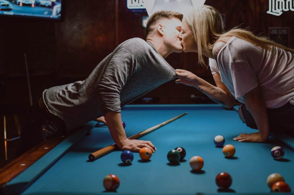 Photo of a blonde woman pulling a blonde man into a kiss over a pool table at a bar.