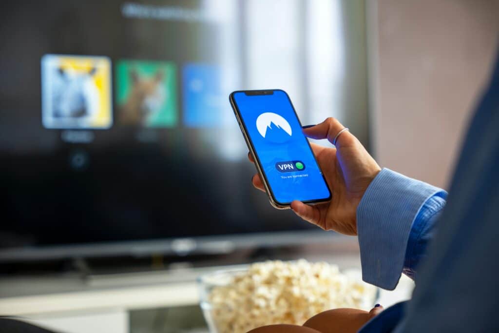 Photo of a person holding a phone displaying a VPN screen, while in the background is a TV displaying a Roku screen and a bowl of popcorn. 