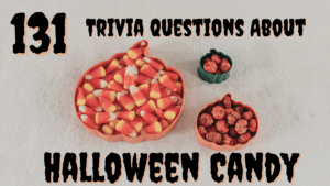 Orange and green pumpkin shaped bowls holding candy corn and pumpkin candies against a beige background, with black and orange text around it that reads "131 Trivia Questions About Halloween Candy"