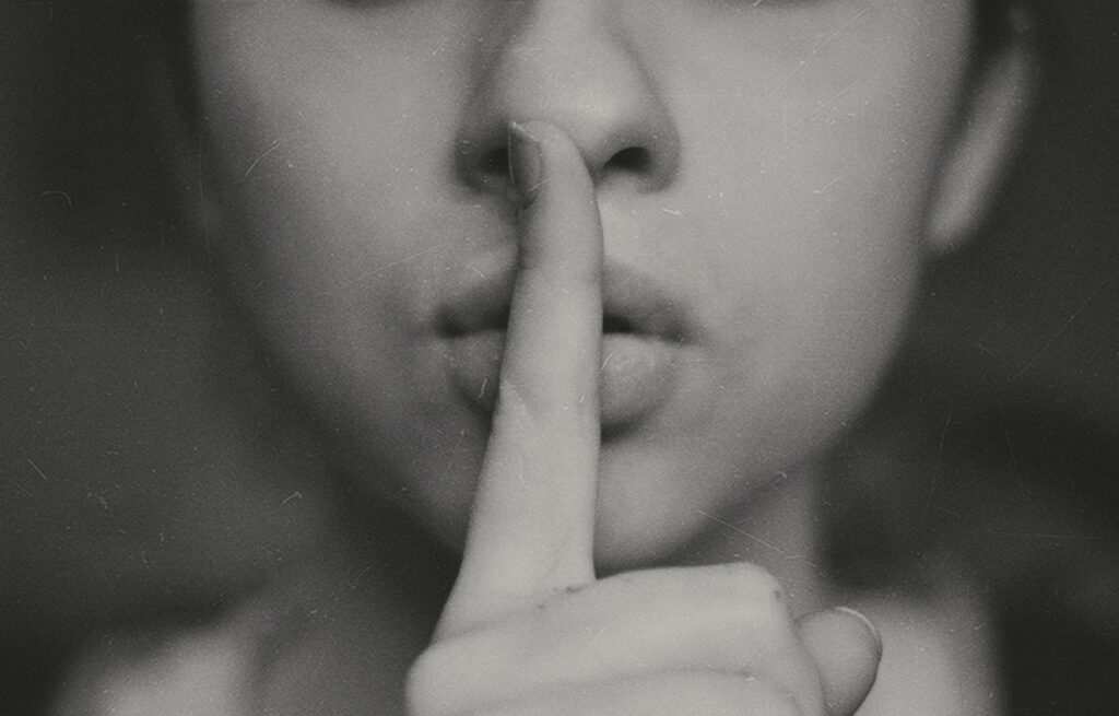 Black and white grainy photo of a woman holding her finger to her lips, signifying silence. 