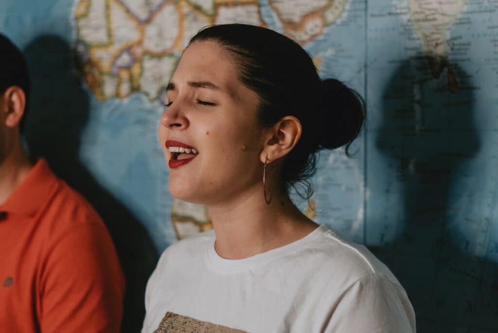 Photo of a brunette woman in a white shirt singing with her eyes closed in front of a map on the wall behind her. 