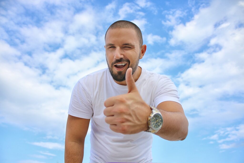 Man in a white shirt wearing a watch and giving a thumbs up in front of a blue sky with white clouds. 