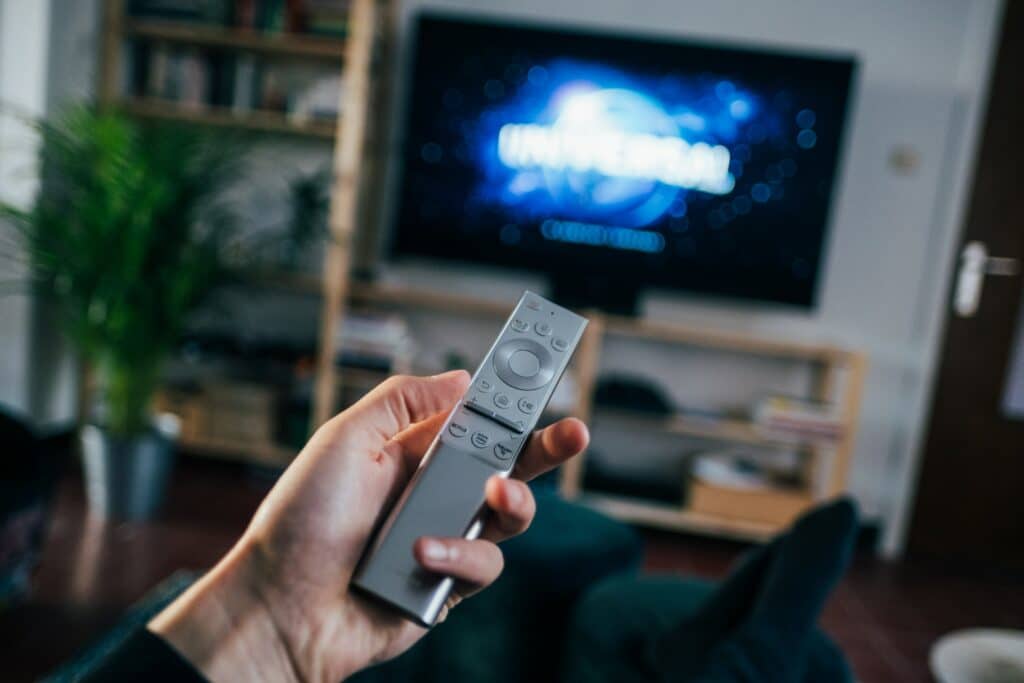 Photo of a person holding a silver remote, a blurry TV in the background is displaying the Universal pictures logo. 