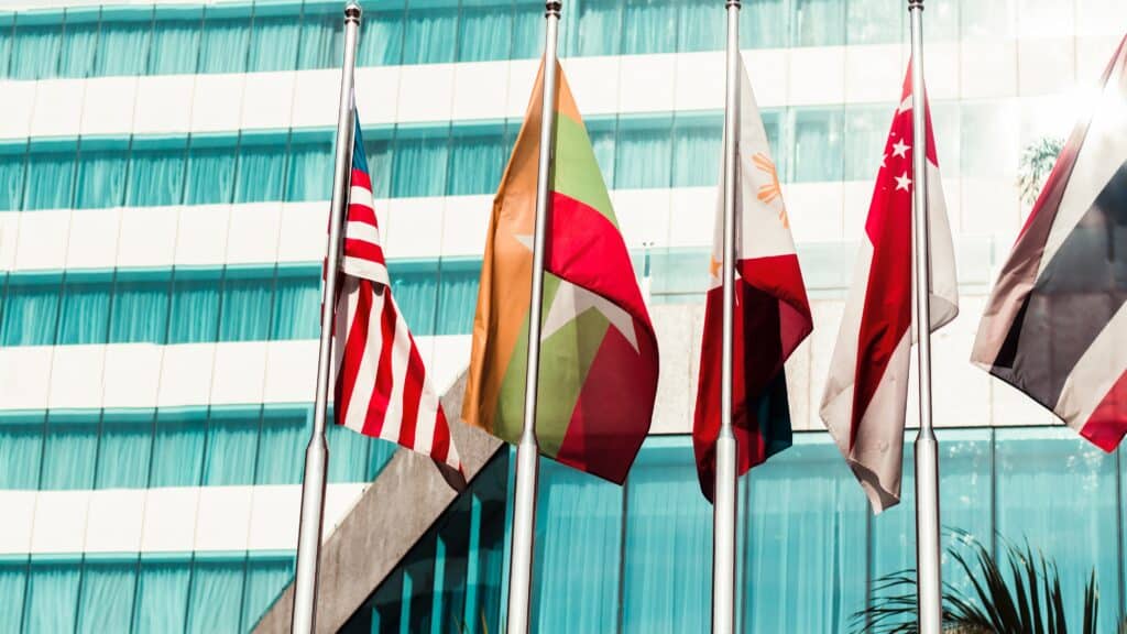 Photo of various flags from around the world on flagpoles outside a glass building.