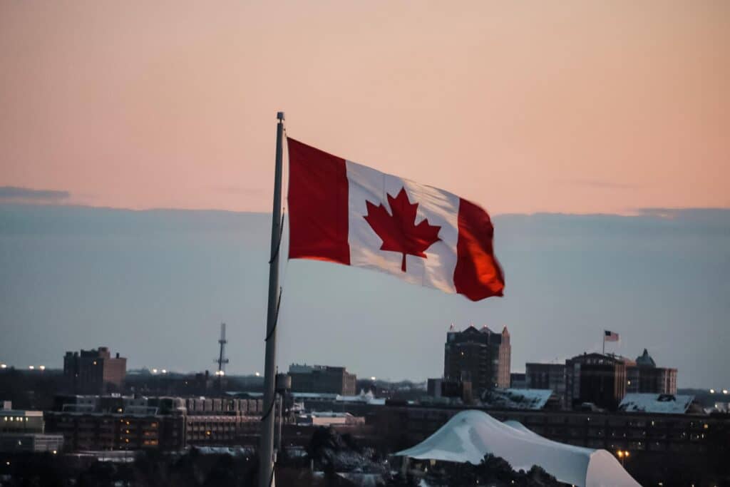 Photo of the Canadian flag flying at dusk with various buildings behind it.