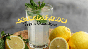 Photo of a glass of lemonade garnished with mint, surrounded by cut and whole lemons on a wooden table against a grey background. Yellow and black text atop it reads "Lemonade Trivia Questions"
