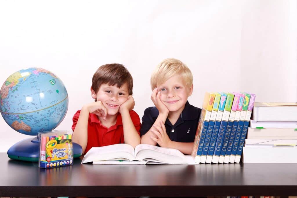 Photo of two young boys, one in a red polo and one in a black polo, posing at a desk with an open book in front of them. On the desk also are a globe, markers, a series of Childcraft books, and a stack of large textbooks.