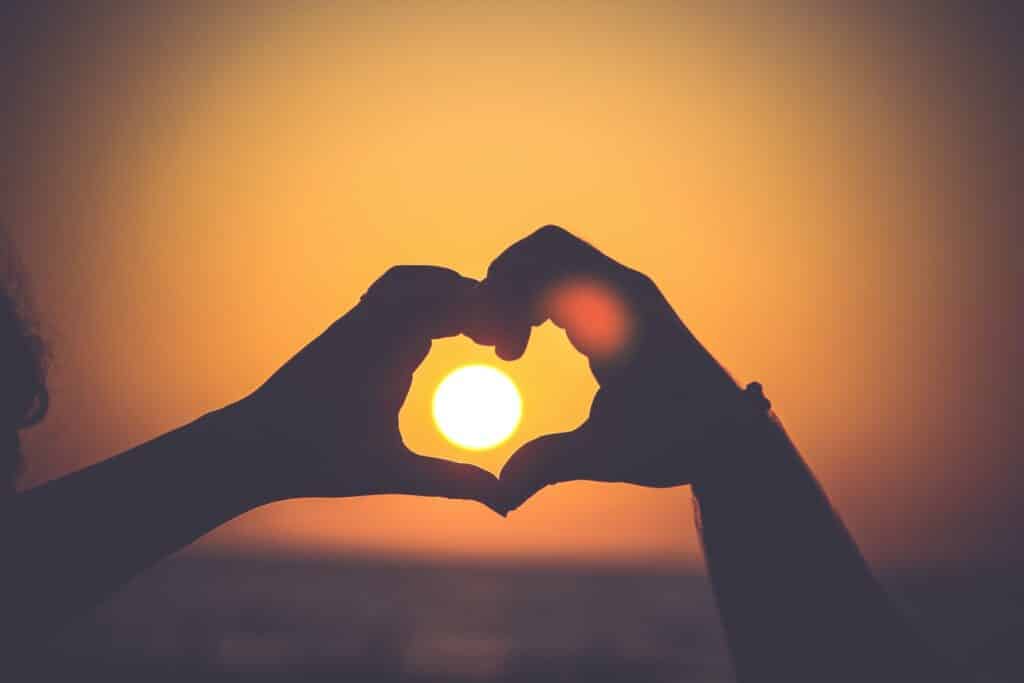 Photo of two hands put together, making a heart, with the sunset shining between them in the background.