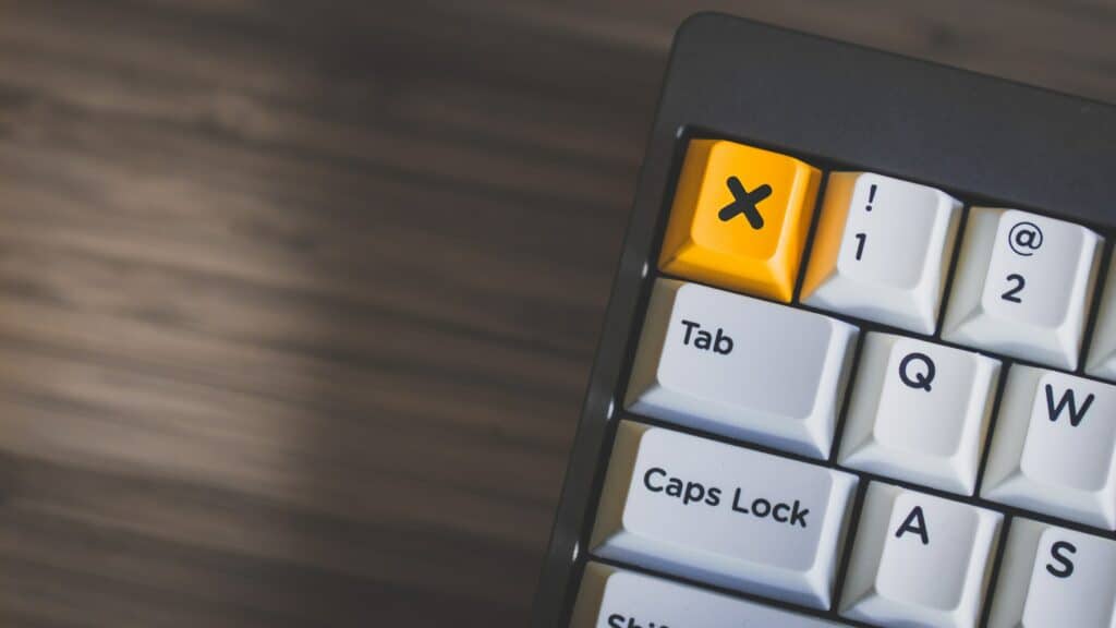 Close up of the corner of a keyboard against a wooden desk. The delete key is yellow with a black X on it. 
