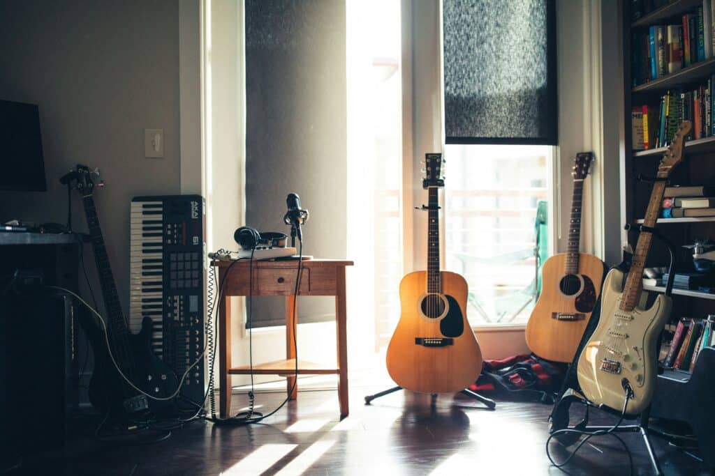Photo of a naturally-lit room with a bookshelf, three guitars, one bass guitar, a keyboard, headphones, and a microphone.